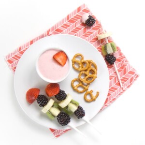 round white plate on pink napkin, small while bowl on plate filled with strawberry cream cheese dip, fruit kababs and a small handful of pretzels