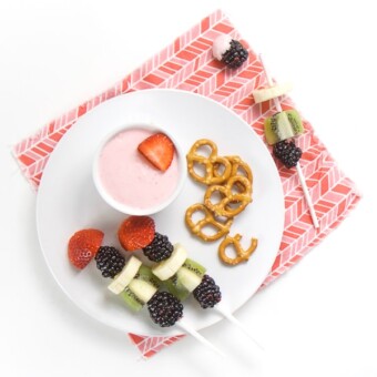 round white plate on pink napkin, small while bowl on plate filled with strawberry cream cheese dip, fruit kababs and a small handful of pretzels