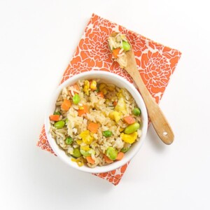 white bowl filled with brown rice, veggies and chunks of scrambled egg. white bowl is sitting on an orange napkin with flowers on it.