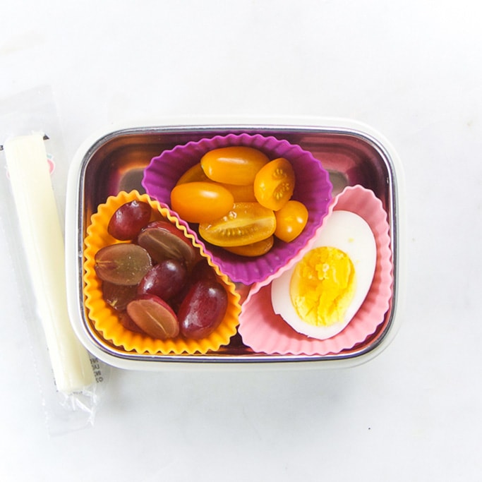 Rectangle kids bento box with healthy snacks for kids - 3 muffin molds filled with cut grapes, cut cherry tomatoes and half a hard boiled egg. On the side is a string cheese. 