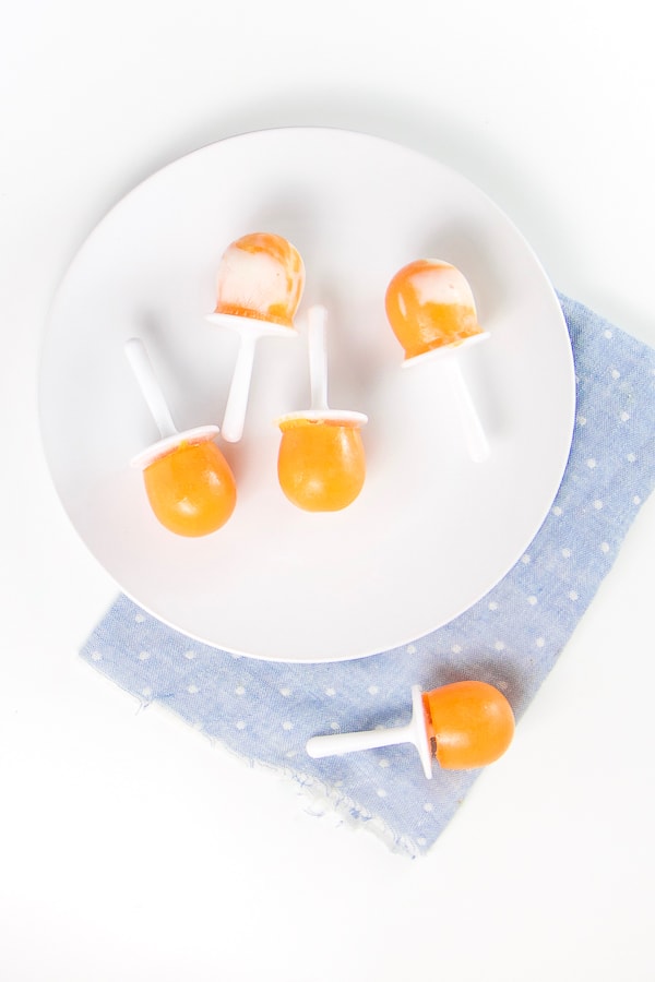 round white plate on top of a blue napkin. On the plate there are 4 orange popsicles with yogurt swirled around in them. there is one orange popsicle on the napkin. 