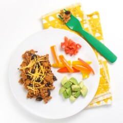 round white plate with a taco meal for baby - finger foods of turkey taco meat with beans and a sprinkle of cheese, on the side - chunks of avocado, strips of peppers and chunks tomatoes.