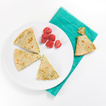 white round plate with wedges of kale pesto quesadilla and a side of chopped raspberries. The plate is sitting on a teal napkin and there is a torn wedge of quesadilla on it.