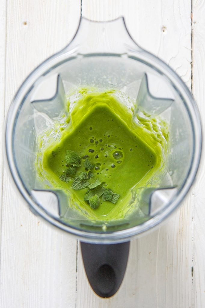 a blender filled with green asparagus puree with mint leaves on top, sitting on a white wooden board
