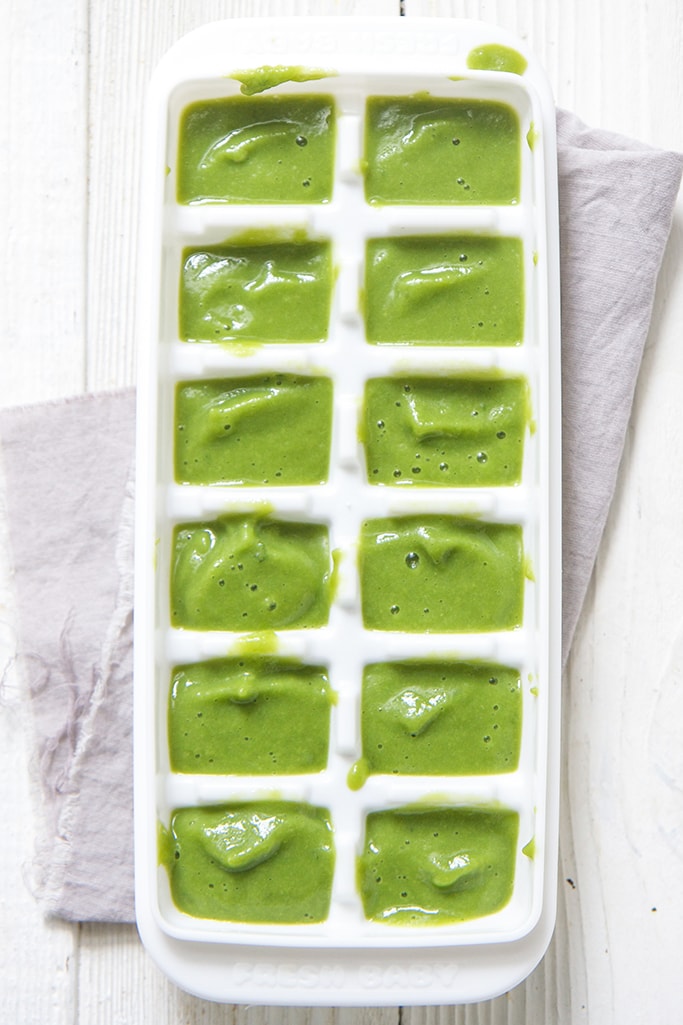 white freezer tray filled with green asparagus baby food sitting on top of a grey napkin on top of a white wooden board. 