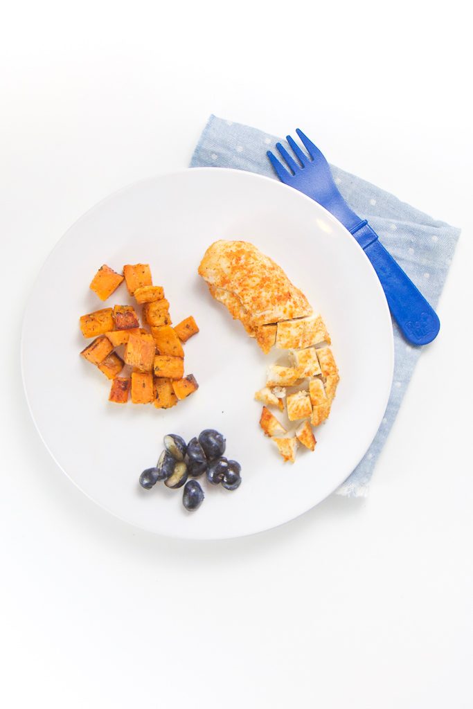 finger foods on a round white plate filled with a chicken tender part way cut into chunks, small chunks of sweet potato and cut blueberries. 