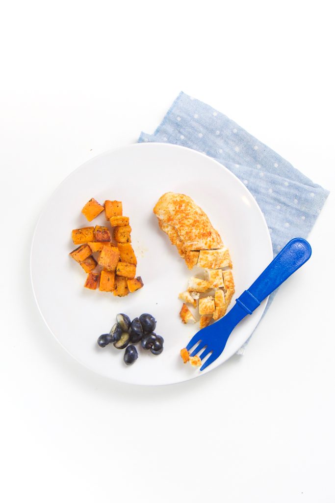 round white plate sitting on top of a blue napkin with chunks of roasted sweet potato, cut blueberries and a chicken tender cut into finger foods for baby. 