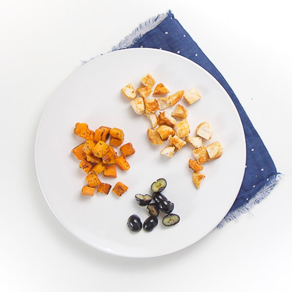 round white plate on top of a blue napkin. On plate is cut up baked chicken, roasted sweet potatoes and cut blueberries.