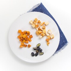 white round plate on top of a dark blue napkin. On the plate is finger foods for baby or toddler - chopped seasoned chicken, blueberries and sweet potatoes