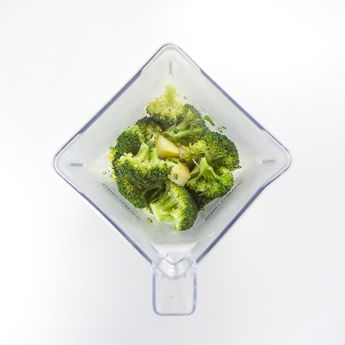 overhead shot of a blender filled with broccoli and chives on a white background