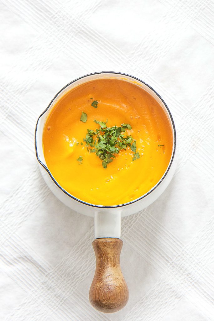 small white saucepan sitting on top of a white tablecloth, inside is a thick butternut squash baby puree with a sprinkle of chopped cilantro on top. 