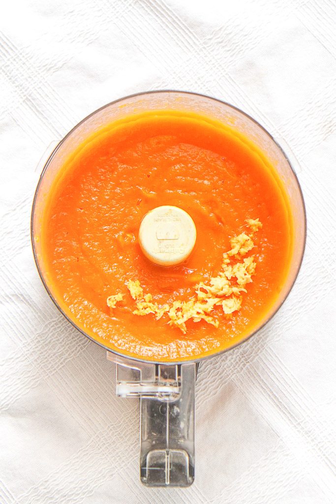 food processor sitting on a white tablecloth filled with carrot and ginger baby food puree with a sprinkle of freshly chopped ginger on top. 