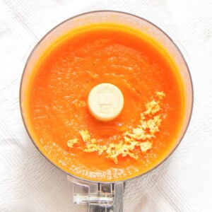 food processor sitting on a white tablecloth filled with carrot and ginger baby food puree with a sprinkle of freshly chopped ginger on top.