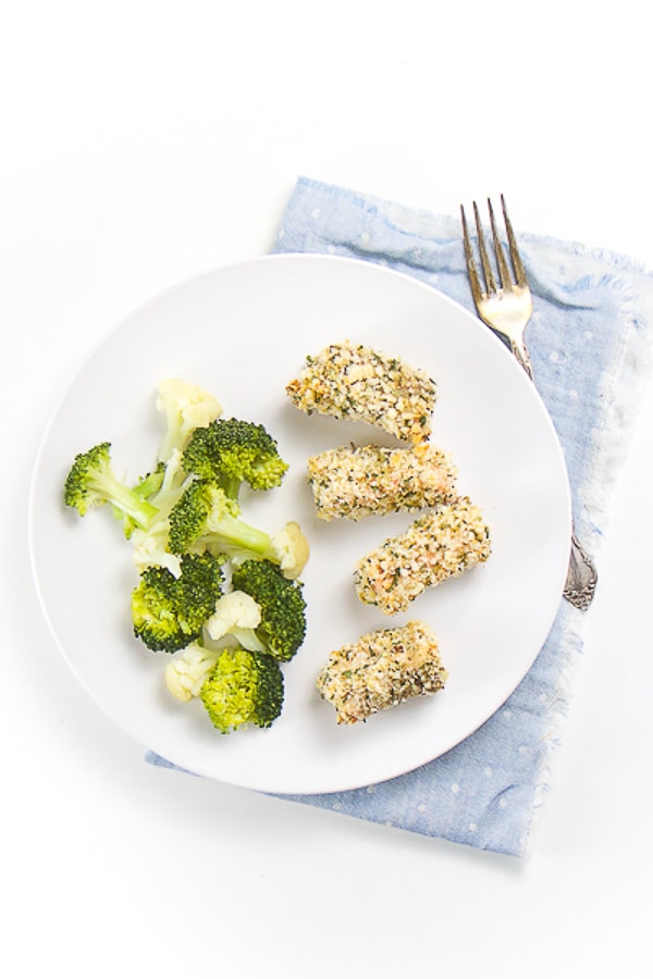 round white plate on top of a blue napkin. On the plate is 4 salmon bites coated in breadcrumbs and herbs. On the side is steamed broccoli and cauliflower.