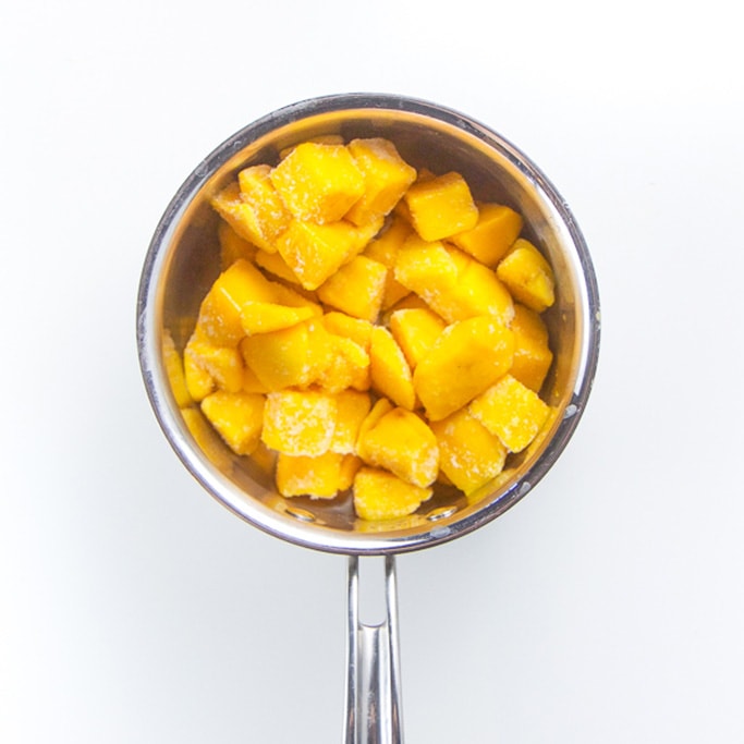silver saucepan sitting on a white background filled with frozen mango chunks