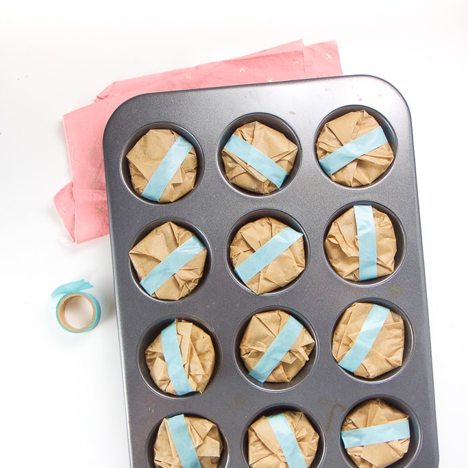 overhead shot of a silver muffin tin with tulip liners with the granola bars inside. All of liners have been folded down towards the center and has been taped with teal washi tape. The muffin tin is sitting on a pink napkin on a white background.
