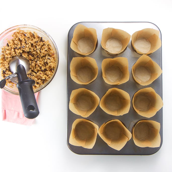 a 12 cup muffin tin is lined with the tulip muffin liners with the bowl of granola mixture next to it.