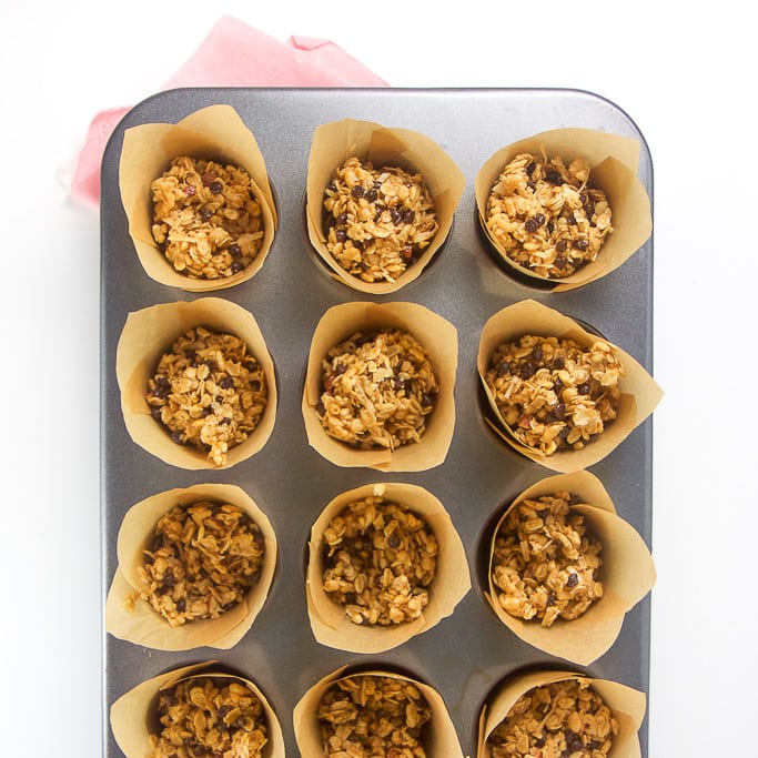 overhead shot of a silver muffin tin with tulip liners with the granola bar mixture inside. The muffin tin is sitting on a pink napkin on a white background.
