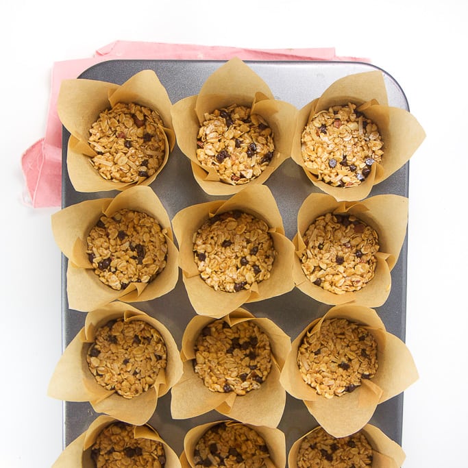 overhead shot of a silver muffin tin with tulip liners with the granola bar mixture pressed down on the inside. The muffin tin is sitting on a pink napkin on a white background.
