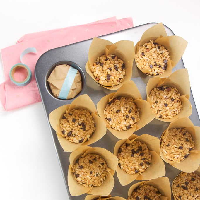 overhead shot of a silver muffin tin with tulip liners with the granola bars inside. One of the liners has been folded down towards the center and has been taped with teal washi tape. The muffin tin is sitting on a pink napkin on a white background.