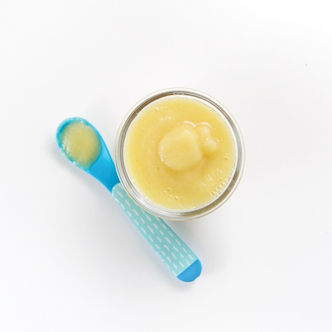 an overhead shot of a small glass jar filled with a creamy apple puree with a blue spoon sitting on the white background filled with baby food puree