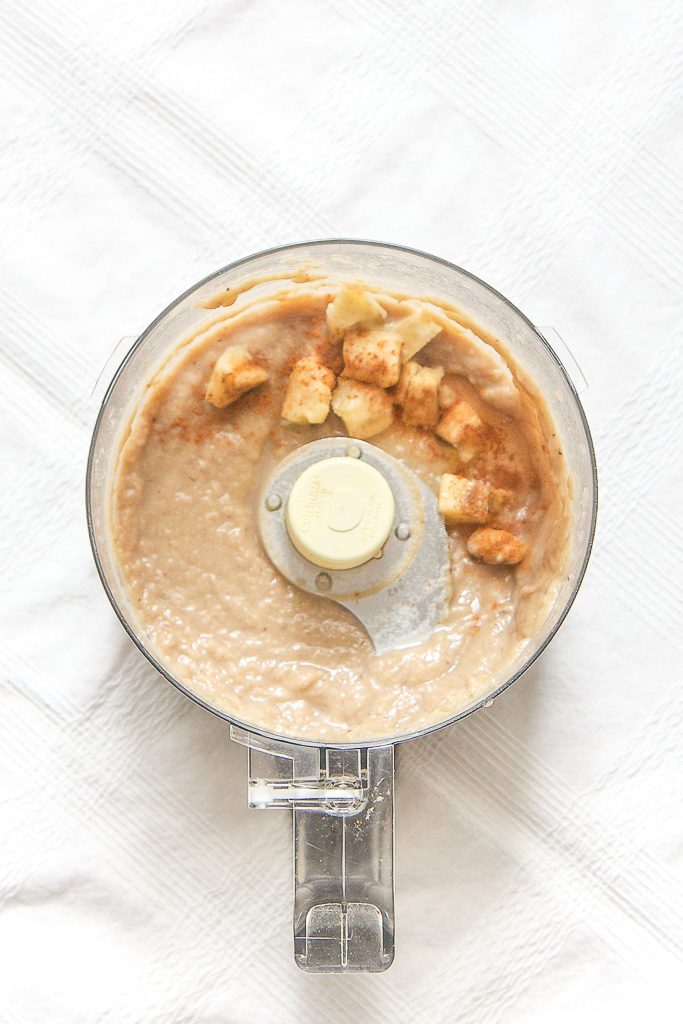 food processor sitting on a white tablecloth filled with a roasted banana baby puree with chunks of bananas and cinnamon sprinkled on top. 