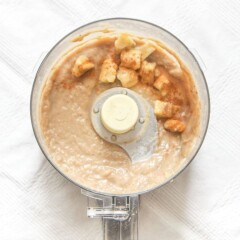 food processor sitting on a white tablecloth filled with a roasted banana baby puree with chunks of bananas and cinnamon sprinkled on top.