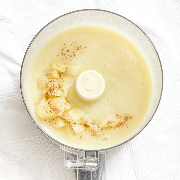 A clear food processor is filled with a pear puree that has chunks of cooked pear and nutmeg sprinkled on top. 