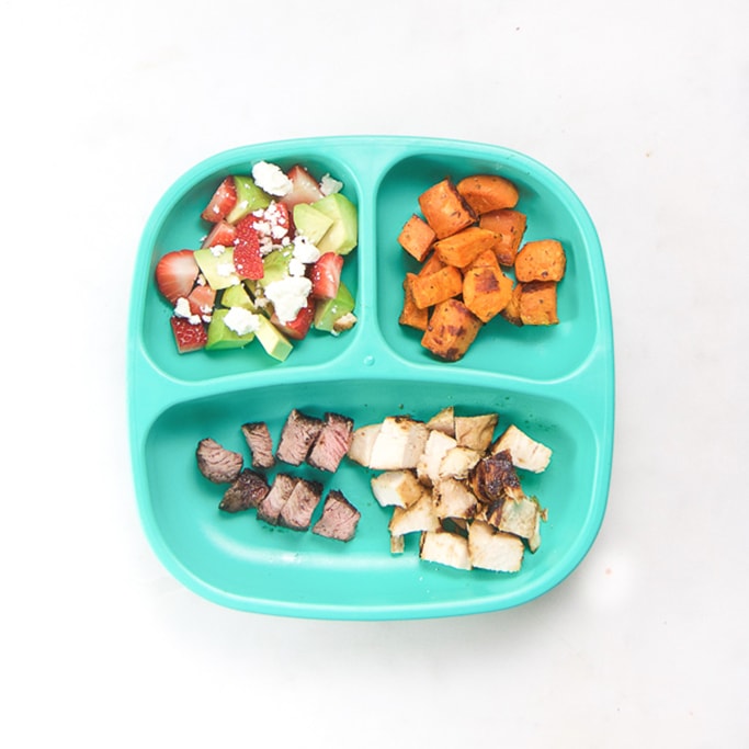 3-section plate on a white surface filled with toddlers dinner meal - cut up steak and chicken, roasted sweet potatoes, strawberries and avocado.  