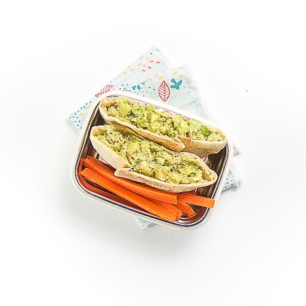 a white bento box is filled with a mini pita pocket cut in half and filled with an avocado tuna salad, there are cut sticks of carrots on the plate as well. The plate is sitting on a multi-colored napkin on a white surface.