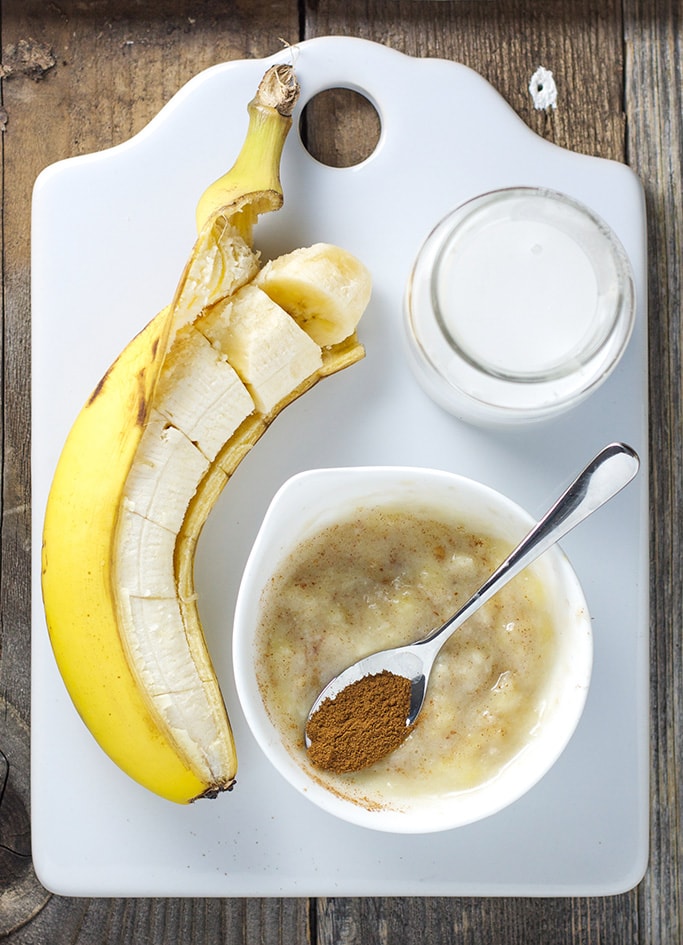 baby food recipe - small round bowl filled with smashed baby food puree with a silver spoon resting inside the bowl filled with cinnamon. On a white background is a banana cut into chunks and small glass of coconut milk.