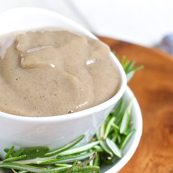 A small white bowl filled with a thick and creamy banana baby food puree, surrounded by fresh rosemary
