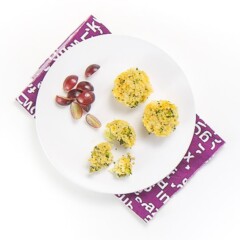 white plate sitting on top of a purple napkin on a white surface. On the plate is 3 mini cheesy broccoli quinoa bites and a small handful of sliced grapes. One of the quinoa bites is halfway eaten with a fork.