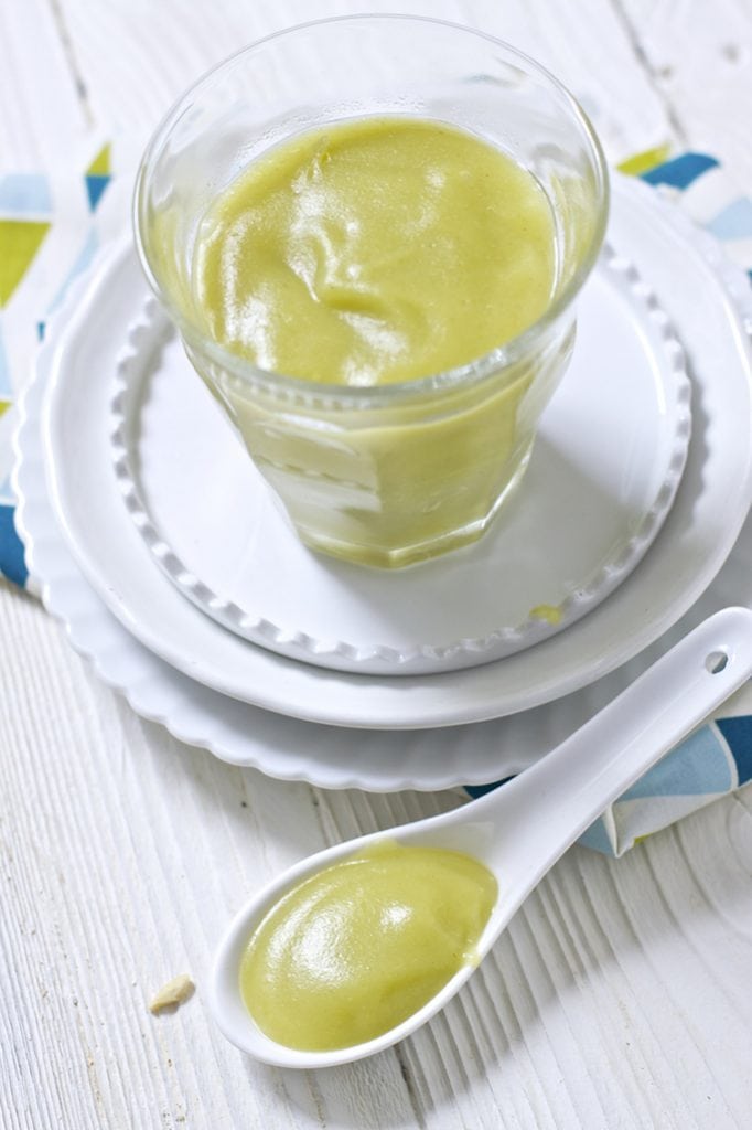 A clear glass jar is sitting on top of 2 white round plates. There is white spoon sitting next to the jar and both the jar and spoon are filled with a leek baby food puree. 