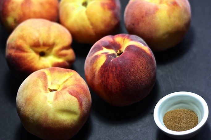 Ripe peaches and a small bowl of cloves sitting on a black cutting board. 