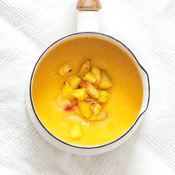 A small white saucepan is filled with a peach puree with chunks of peaches and vanilla beans sprinkled on top