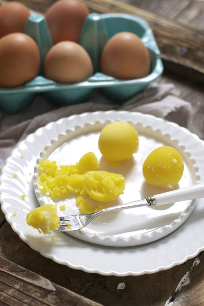 white plate with 3 hard boiled egg yolks with a fork cutting one apart. There is a teal container of eggs behind the plates and everything is on a rustic wood surface. 