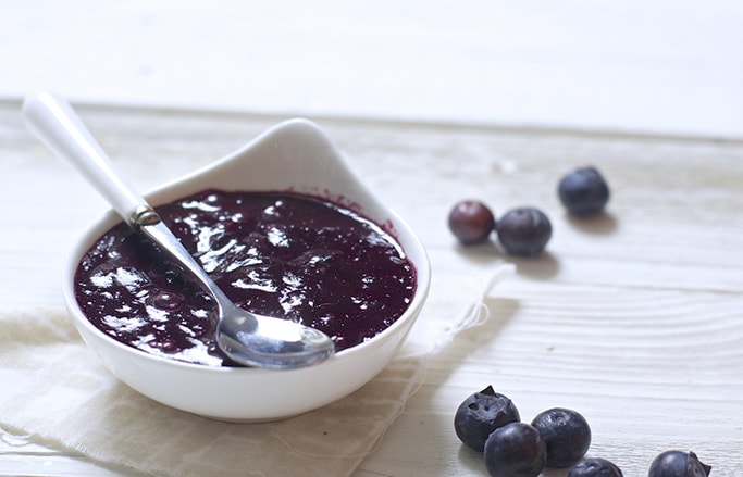 small white bowl filled with a blueberry baby food puree inside. bowl has a white and silver spoon resting on top and is sitting on a white surface