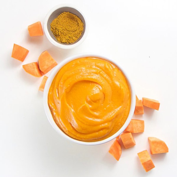 A small white bowl is filled with sweet potato puree has has chunks of sweet potatoes and a small bowl of mild curry are scattered around it. 
