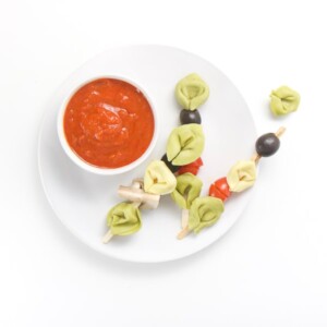 small white plate sitting on a white surface. On top of plate is a small white bowl filled with a dipping sauce and 3 wooden sticks filled with tortellini and veggies, a couple of tortellini and veggies are scattered on the table.