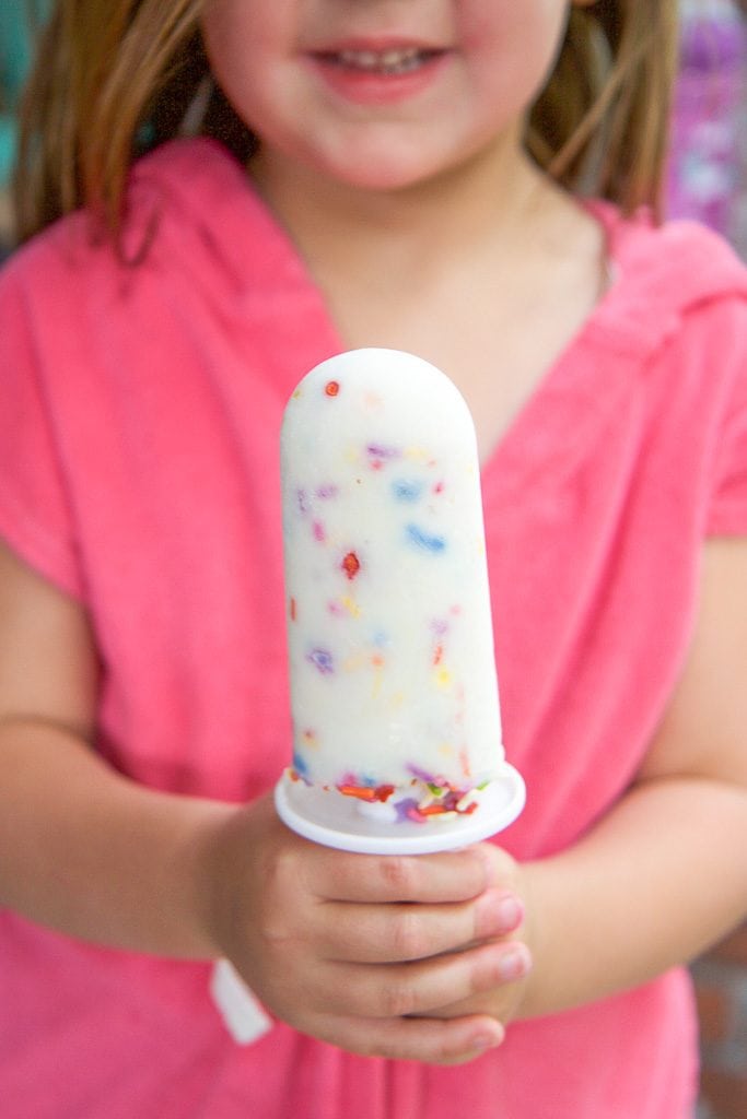 A small girl wearing pink holding a healthy funfetti popsicle out in front of her. 