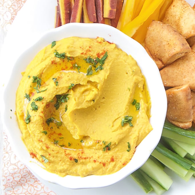 A white bowl filled with carrot hummus with veggies and pita around it. 
