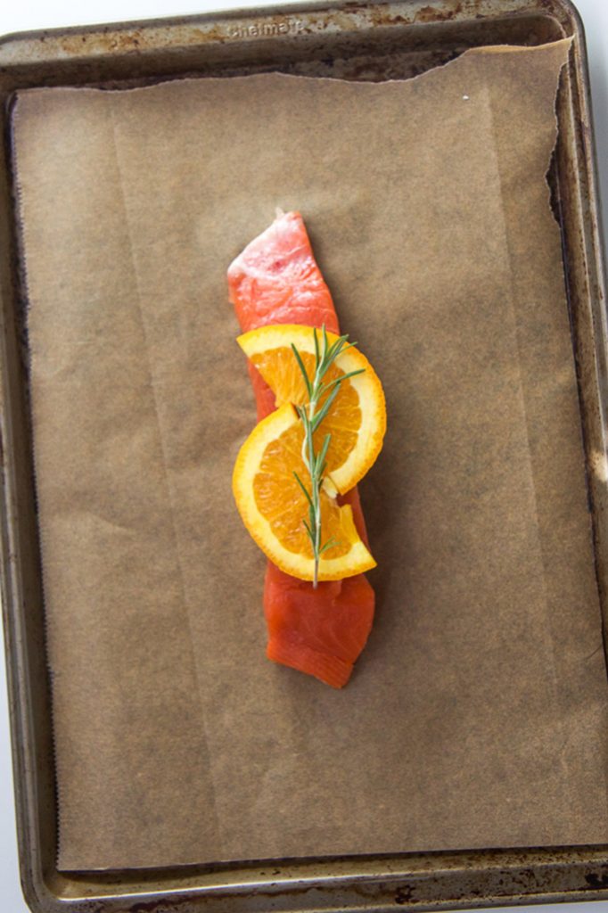 A piece of salmon sitting on a parchment lined baking sheet with orange and rosemary on top. 
