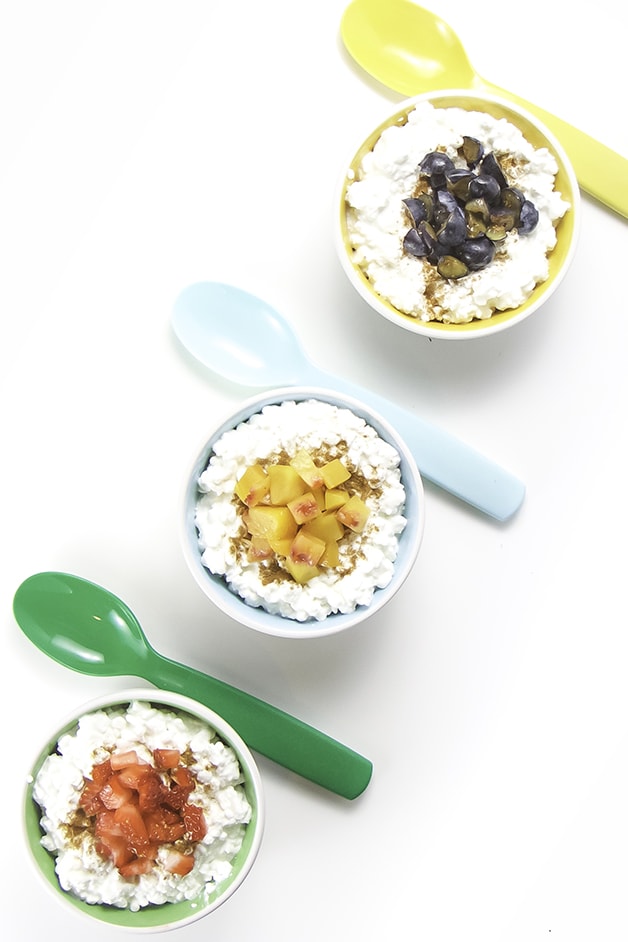 3 small bowls filled with cottage cheese an topped with chunks of fruit are lined up with spoons resting next to each bowl. 