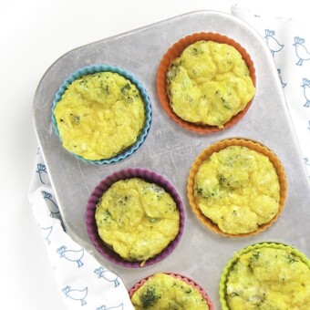 A silver muffin tin filled with broccoli egg cups with one cup on the white surface.