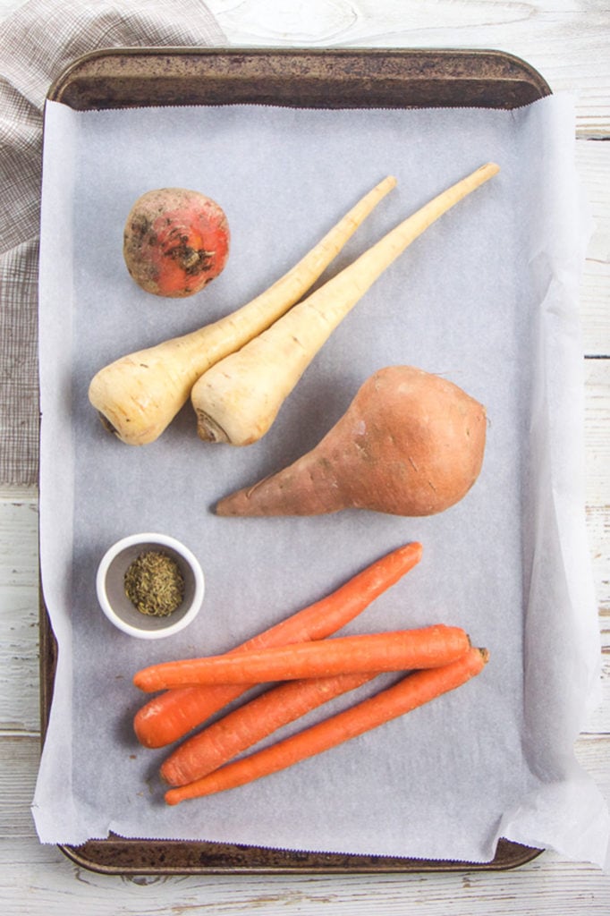 A baking sheet with 4 root veggies on top and a small bowl filled with herbs. 