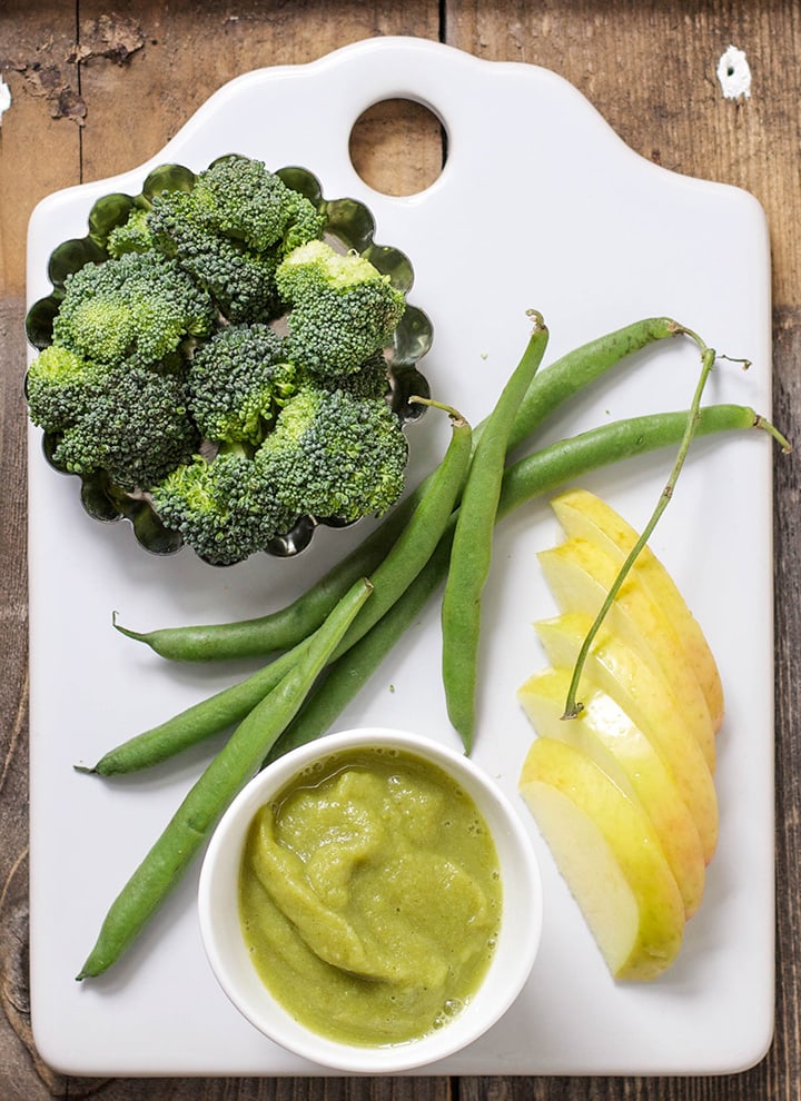 A white cutting board filled with a spread of produce and a small bowl of apple green bean and broccoli baby food puree. 