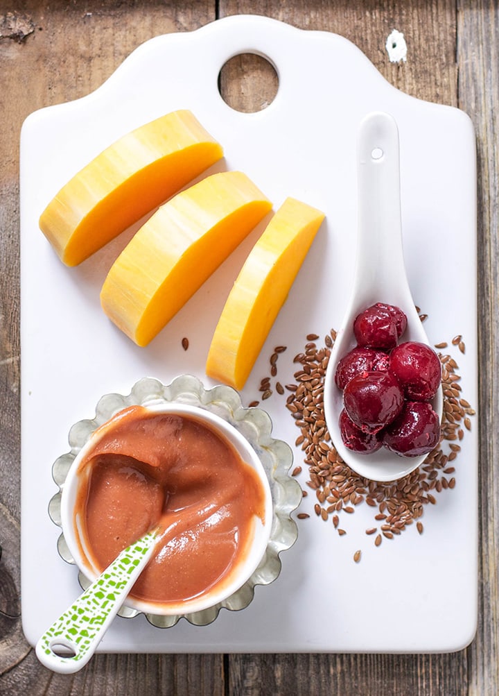 A white cutting board with chopped butternut squash, cherries, flax seeds and a baby food puree in a small white bowl. 