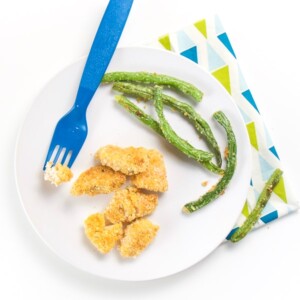 Round white plate with chicken nuggets and green beans on top. Fork resting on place with chicken on it.