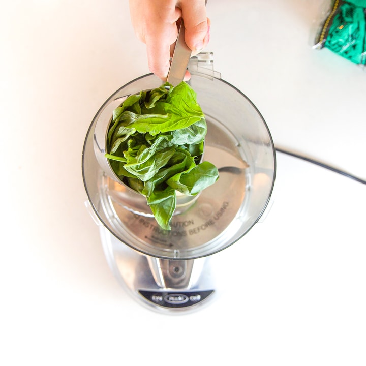 Hand reaching out with a cup full of basil about to dump it into a food processor.